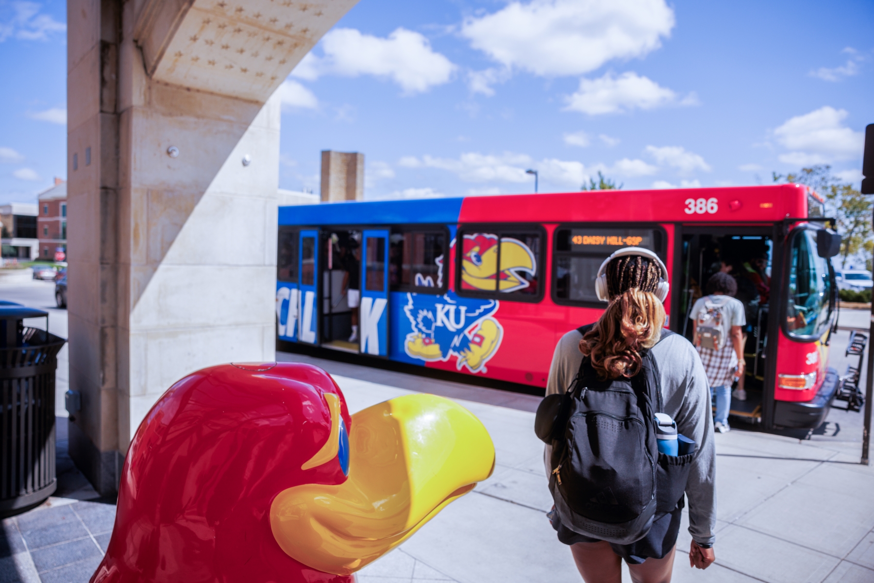A University of Kansas bus, with students walking toward it from the Union. Jayhawk statue looks over the campus from the bottom left of image. 