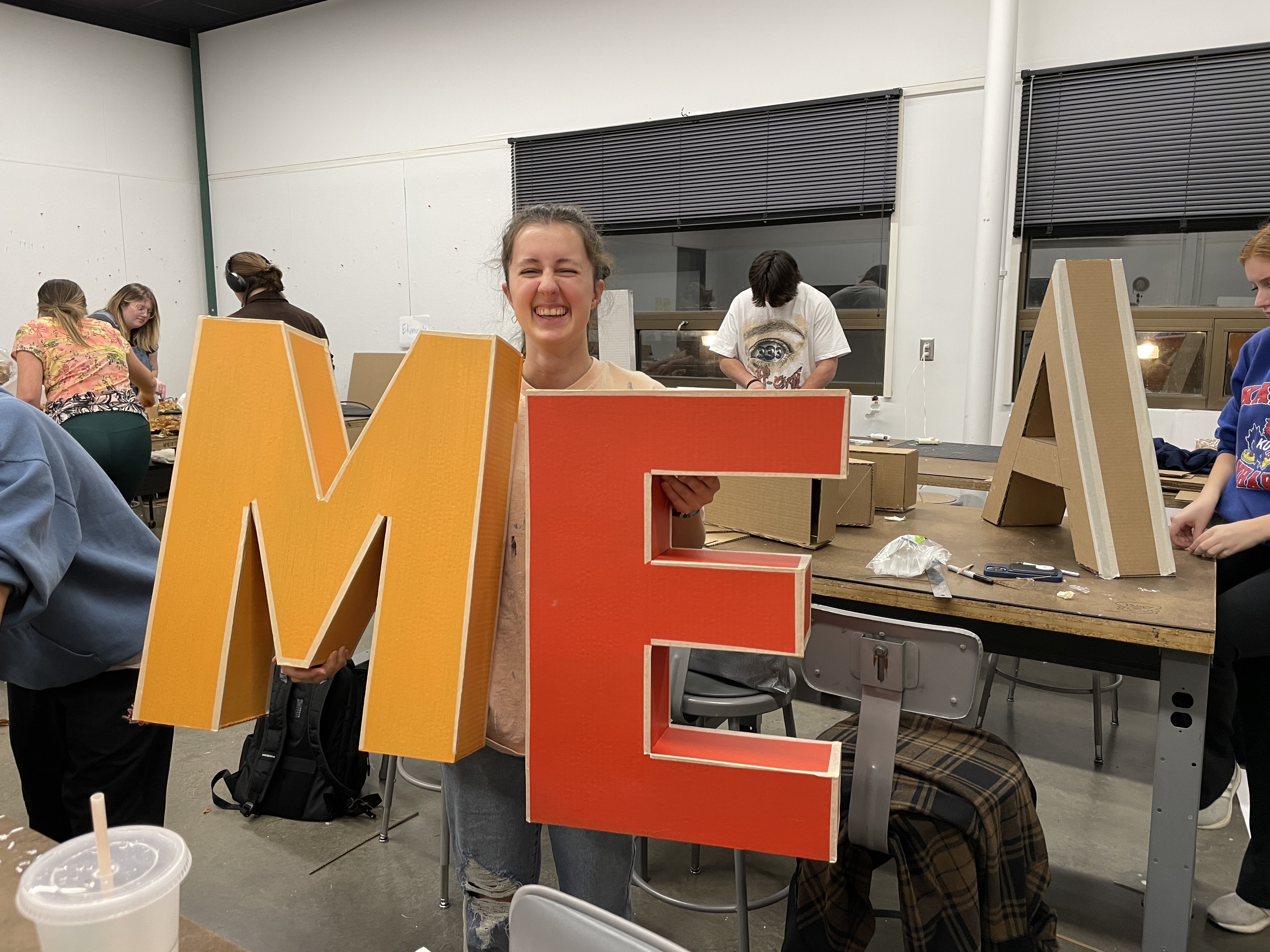 A KU industrial design student holding up large orange and red letters "M" and "E" in a classroom setting. 