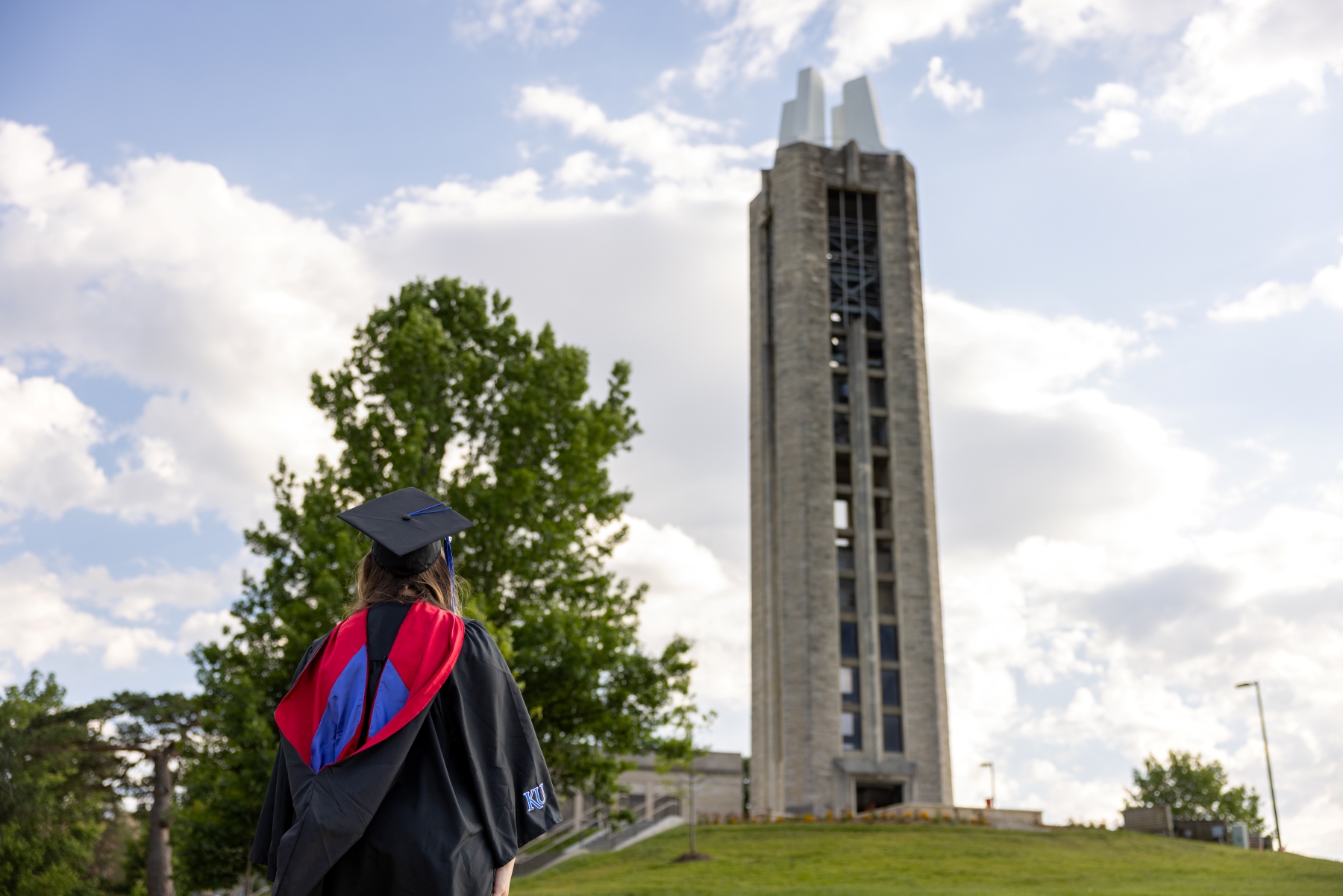A woman in a University of Kansas graduate degree regalia stands on the Hill beneath the Campanile.
