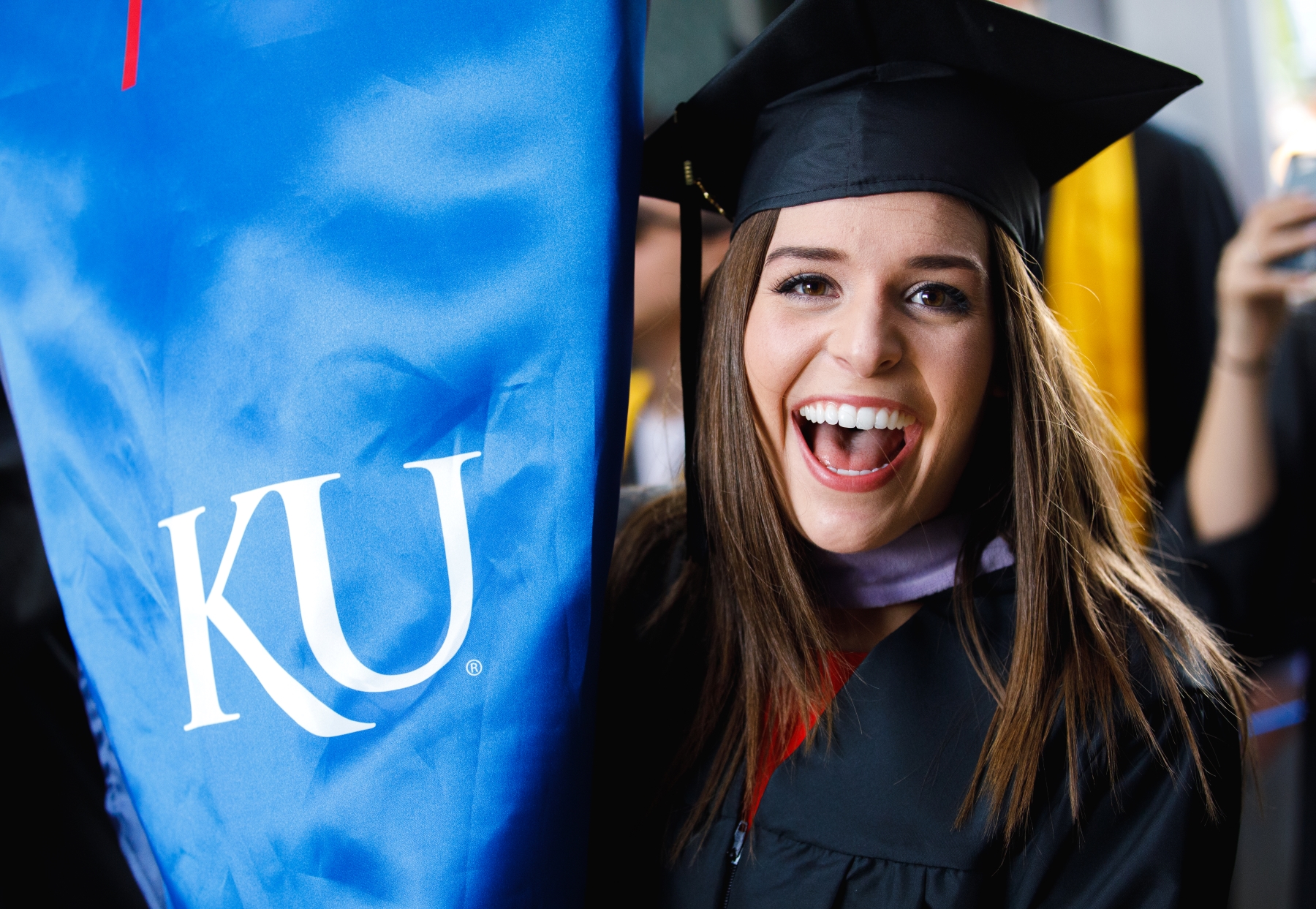 A woman smiles at the camera next to her University of Kansas graduation regalia.