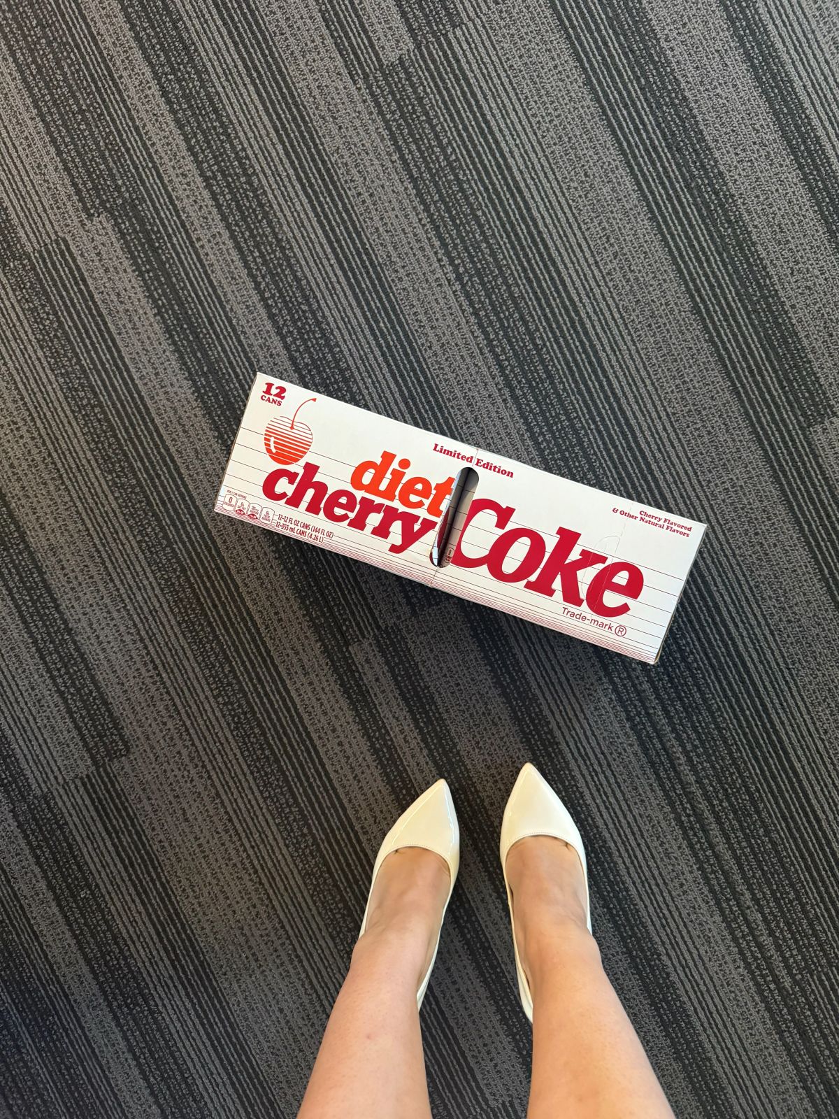 An image of a 12 pack of Diet Cherry Coke, next to an unidentified woman's pair of white heels.